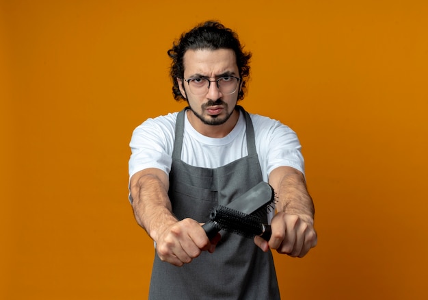 Fruncir el ceño joven barbero masculino caucásico con gafas y banda de pelo ondulado en uniforme estirando peines hacia la cámara aislada sobre fondo naranja con espacio de copia