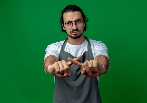 Foto gratuita fruncir el ceño joven barbero masculino caucásico con uniforme y gafas sin hacer ningún gesto a la cámara aislada sobre fondo verde con espacio de copia