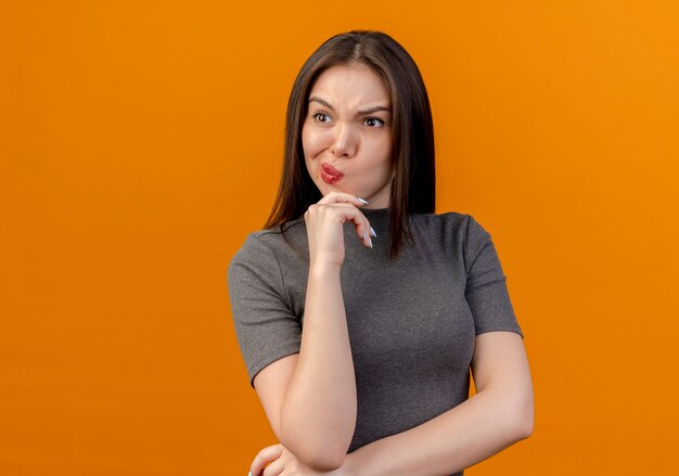 Fruncir el ceño joven bonita mujer poniendo las manos debajo del codo y en la barbilla mirando al lado aislado sobre fondo naranja con espacio de copia