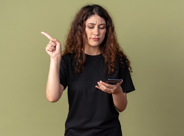 Fruncir el ceño joven bonita sosteniendo y mirando el teléfono móvil apuntando al lado