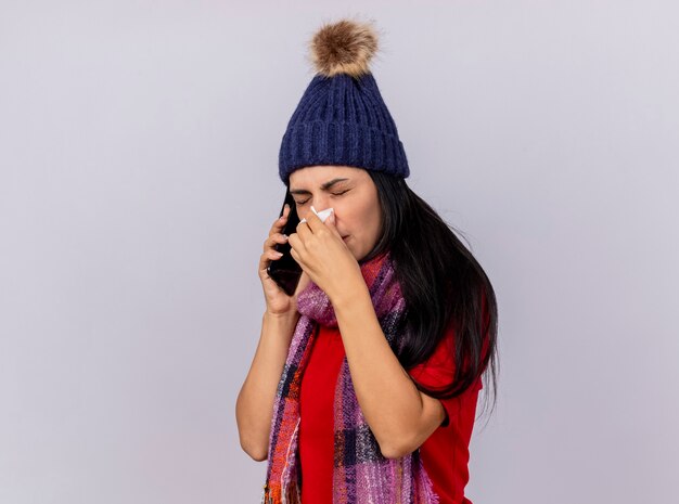 Fruncir el ceño a joven caucásica enferma con sombrero de invierno y bufanda de pie en vista de perfil hablando por teléfono limpiando la nariz con una servilleta aislado sobre fondo blanco con espacio de copia