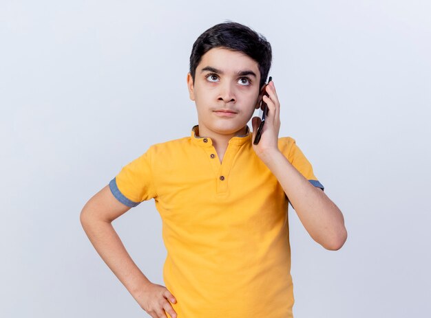 Fruncir el ceño joven caucásico hablando por teléfono manteniendo la mano en la cintura mirando hacia arriba aislado sobre fondo blanco.