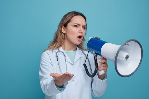 Fruncir el ceño joven doctora vistiendo bata médica y un estetoscopio alrededor del cuello mirando al lado apuntando al lado con la mano hablando por el orador