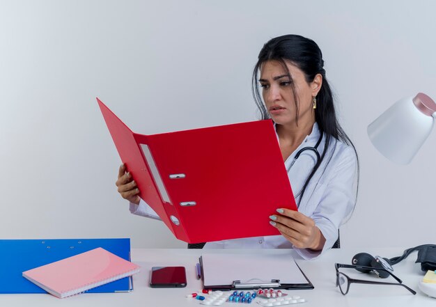 Fruncir el ceño joven doctora vistiendo bata médica y estetoscopio sentado en el escritorio con herramientas médicas sosteniendo y mirando la carpeta aislada