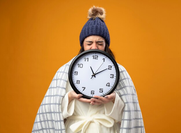 Fruncir el ceño joven enferma vistiendo bata sombrero de invierno envuelto en cuadros sosteniendo el reloj con los ojos cerrados aislado en la pared naranja