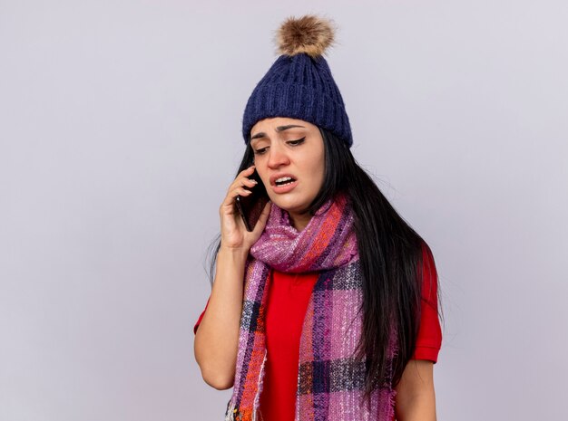 Fruncir el ceño joven enferma vistiendo gorro y bufanda de invierno hablando por teléfono mirando hacia abajo aislado en la pared blanca