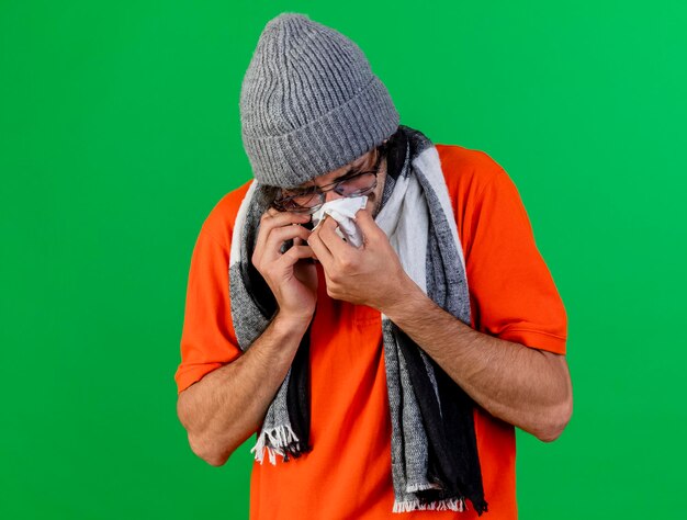 Fruncir el ceño a joven enfermo con gafas, gorro de invierno y bufanda hablando por teléfono limpiando la nariz con una servilleta con los ojos cerrados aislados en la pared verde con espacio de copia