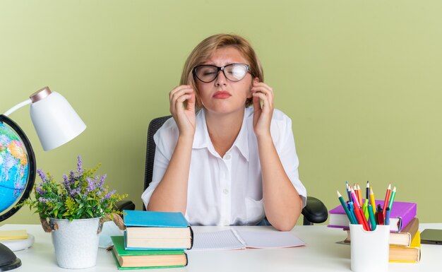 Fruncir el ceño joven estudiante rubia con gafas sentado en el escritorio con herramientas escolares manteniendo las manos en el aire cerca de la cara con los ojos cerrados aislados en la pared verde oliva