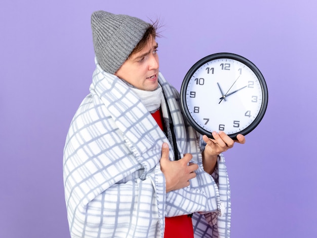 Foto gratuita fruncir el ceño joven guapo rubio enfermo con sombrero de invierno y bufanda envuelto en cuadros sosteniendo y mirando el reloj aislado sobre fondo púrpura