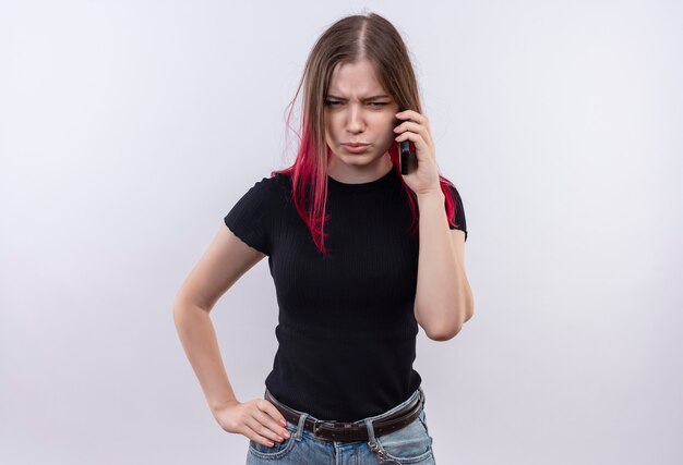 Fruncir el ceño joven hermosa mujer vestida con camiseta negra habla por teléfono poniendo su mano en la cadera en la pared blanca aislada