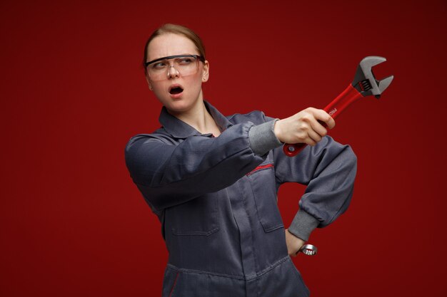 Fruncir el ceño joven ingeniera rubia vestida con uniforme y gafas de seguridad sosteniendo llaves manteniendo la mano en la cintura mirando y apuntando al lado con una llave