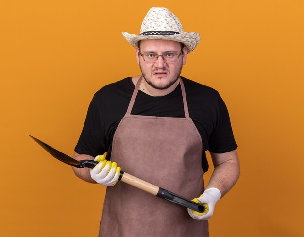 Fruncir el ceño joven jardinero con sombrero y guantes de jardinería sosteniendo la pala aislado en la pared naranja