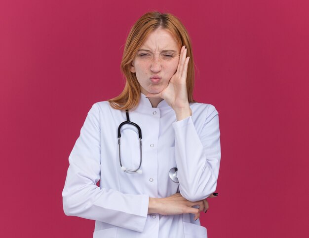 Fruncir el ceño joven médico de jengibre con bata médica y un estetoscopio manteniendo la mano en la cara mirando al frente con los labios fruncidos aislados en la pared carmesí con espacio de copia