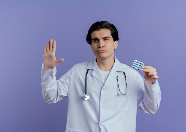 Fruncir el ceño joven médico vistiendo bata médica y un estetoscopio que muestra el paquete de cápsulas haciendo gesto de parada aislado en la pared púrpura con espacio de copia