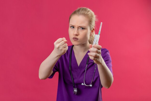 Fruncir el ceño joven médico vistiendo una bata médica púrpura y un estetoscopio sosteniendo la jeringa con el puño levantado en la pared rosa aislada