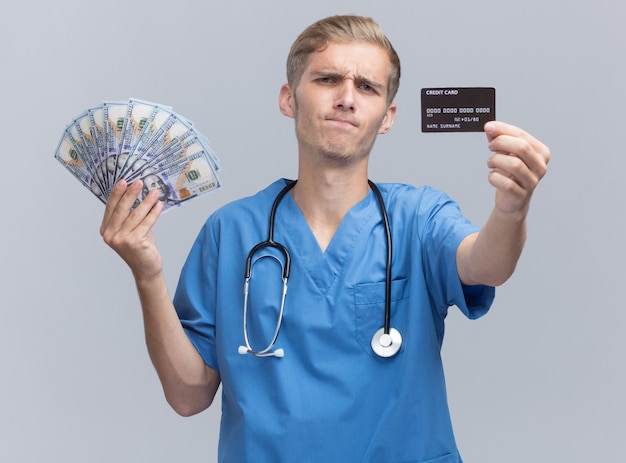 Fruncir el ceño joven médico vistiendo uniforme médico con estetoscopio sosteniendo dinero en efectivo y tarjeta de crédito en la cámara aislada en la pared blanca