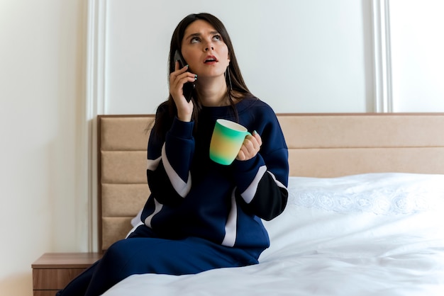 Fruncir el ceño joven mujer bonita caucásica sentada en la cama en el dormitorio sosteniendo la taza mirando hacia arriba y hablando por teléfono