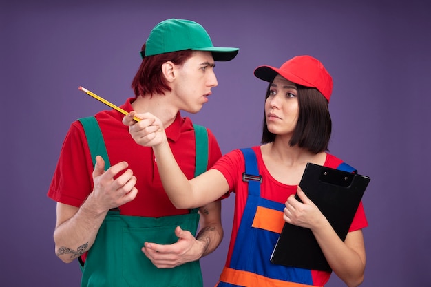 Fruncir el ceño joven pareja en uniforme de trabajador de la construcción y gorra chica sosteniendo lápiz y portapapeles mirando y apuntando al lado con lápiz chico manteniendo la mano en el aire mirando a chica aislada