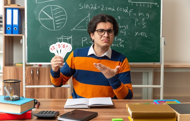 Fruncir el ceño joven profesor de geometría con gafas sentado en un escritorio con útiles escolares en el aula mirando al frente mostrando números de ventiladores apuntando a ellos