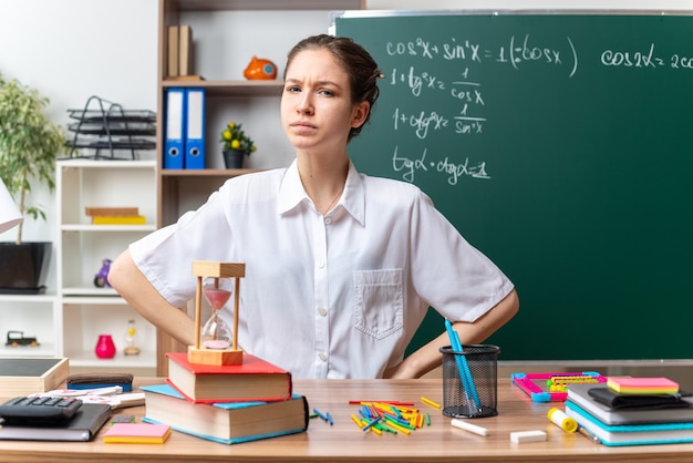 Fruncir el ceño joven profesora de matemáticas sentada en un escritorio con útiles escolares manteniendo las manos en la cintura mirando al frente en el aula