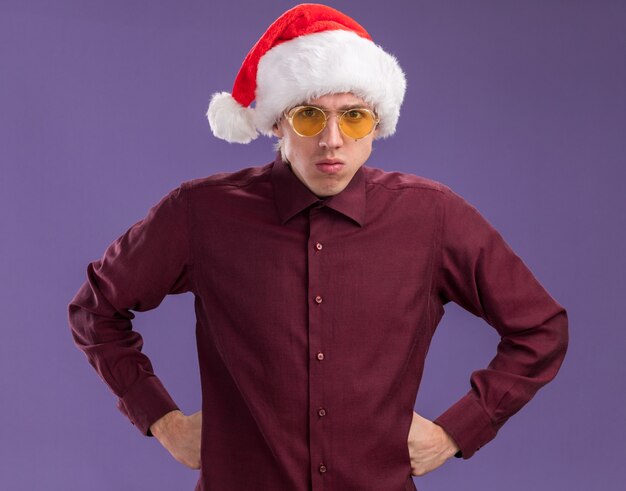 Fruncir el ceño joven rubio con gorro de Papá Noel y gafas manteniendo las manos en la cintura mirando a cámara aislada sobre fondo púrpura