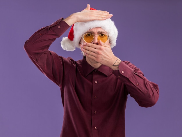 Fruncir el ceño a joven rubio con gorro de Papá Noel y gafas mirando a la cámara manteniendo las manos en la cabeza y en la boca aislada sobre fondo púrpura