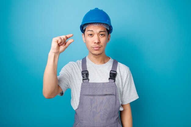 Fruncir el ceño joven trabajador de la construcción con casco de seguridad y uniforme haciendo gesto de pequeña cantidad