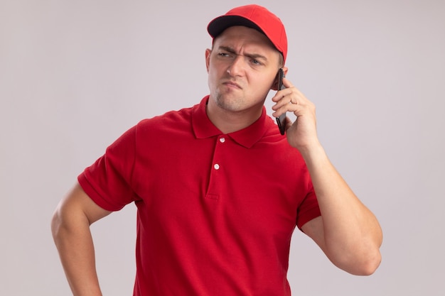 Fruncir el ceño mirando al lado joven repartidor vestido con uniforme con gorra habla por teléfono aislado en la pared blanca