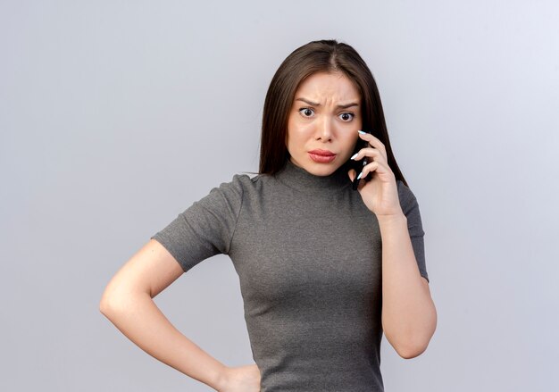 Fruncir el ceño a una mujer bonita joven poniendo la mano en la cintura hablando por teléfono mirando directamente aislado sobre fondo blanco con espacio de copia