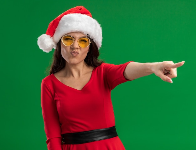 Foto gratuita fruncir el ceño niña bonita con gorro de papá noel y gafas mirando y apuntando al lado aislado sobre fondo verde