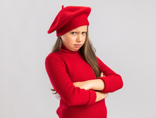 Fruncir el ceño niña rubia vistiendo boina roja manteniendo los brazos cruzados mirando al frente aislado en la pared blanca con espacio de copia