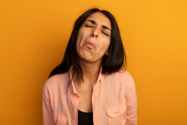 Fruncir el ceño con los ojos cerrados hermosa joven vestida con camiseta rosa aislada en la pared amarilla