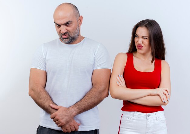 Fruncir el ceño par adulto hombre manteniendo las manos juntas mirando y mujer de pie con postura cerrada mirándolo con un ojo cerrado