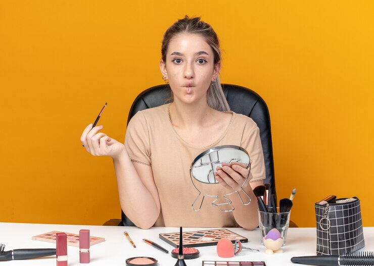 Foto gratuito fruncir los labios hermosa joven se sienta a la mesa con herramientas de maquillaje con pincel de maquillaje con espejo aislado sobre fondo naranja
