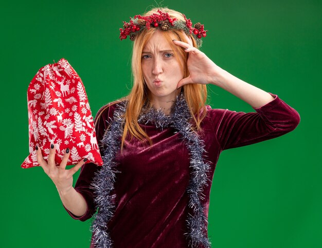 Fruncir los labios joven hermosa chica con vestido rojo con corona y guirnalda en el cuello sosteniendo una bolsa de Navidad poniendo la mano en la cabeza aislada sobre fondo verde