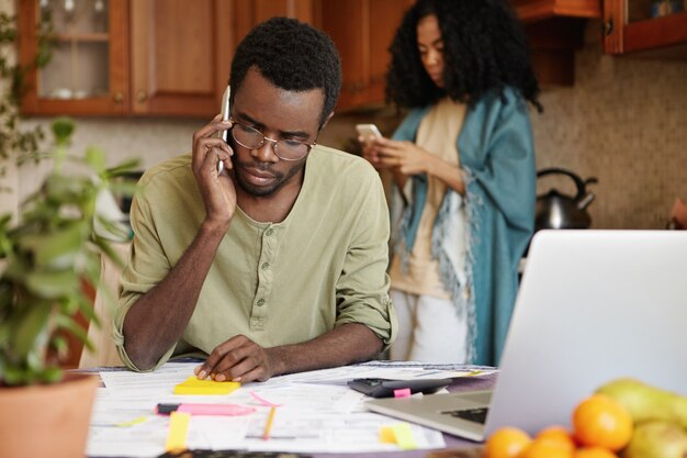 Frustrado joven africano desempleado hablando por teléfono móvil con su amigo, pidiéndole dinero para cubrir los gastos familiares, sin poder pagar más facturas de servicios públicos porque lo despidieron