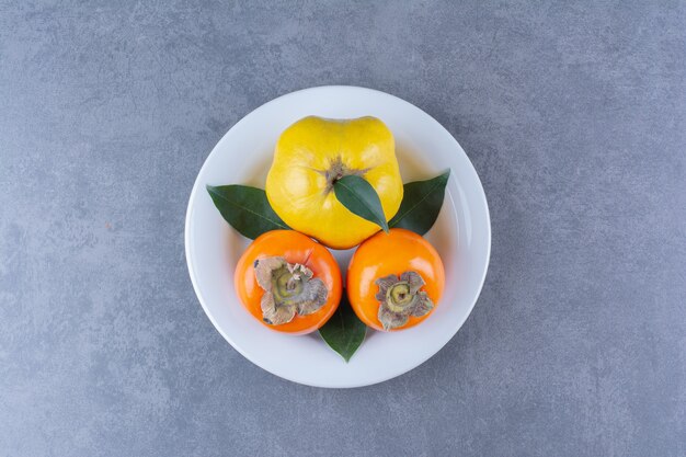 Frutas de membrillo y caqui en placa sobre mesa de mármol.