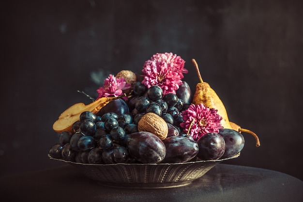 El frutero con uvas y ciruelas contra una pared oscura