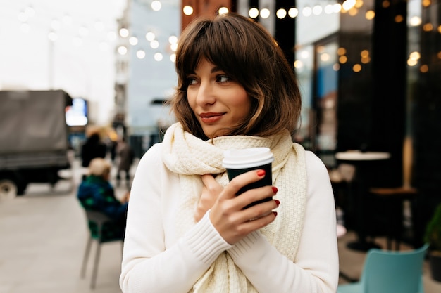 Fuera retrato de encantadora dama bonita con cabello castaño vistiendo un suéter blanco y bufanda bebiendo café en la calle con luces