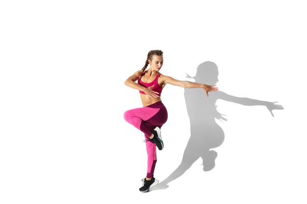 Fuerte. Hermosa joven atleta practicando en la pared blanca, retrato con sombras. Modelo de ajuste deportivo en movimiento y acción. Culturismo, estilo de vida saludable, concepto de estilo.