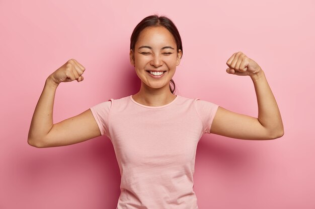 Fuerte y poderosa mujer asiática con cabello oscuro peinado, sonrisa con dientes, levanta los brazos y muestra los bíceps, tiene un piercing en la oreja, usa una camiseta rosada casual, modelos contra la pared rosa. ¡Mira mis músculos!