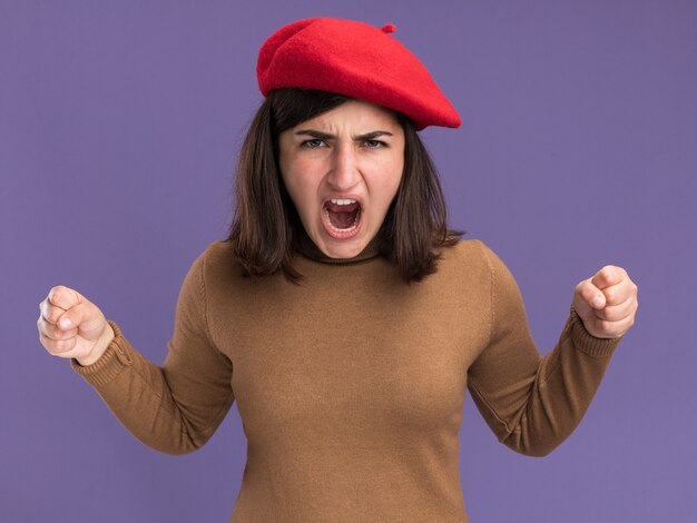 Furiosa joven bastante caucásica con sombrero de boina manteniendo los puños aislados en la pared púrpura con espacio de copia