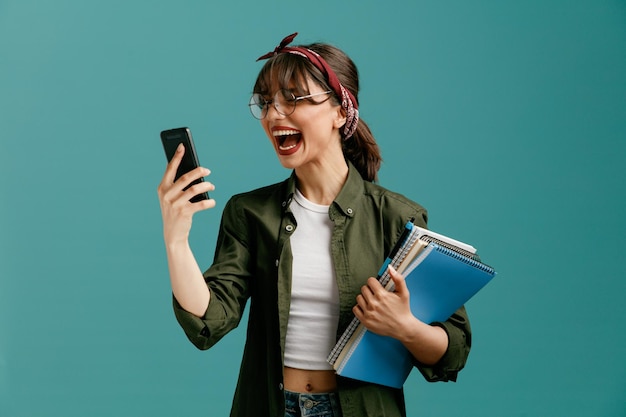 Furiosa joven estudiante con gafas de bandana sosteniendo grandes blocs de notas con bolígrafo y teléfono móvil en otra mano mirando el teléfono móvil gritando en voz alta aislado en el fondo azul