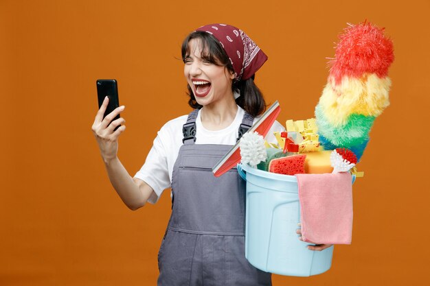 Furiosa joven limpiadora con uniforme y pañuelo sosteniendo un cubo de herramientas de limpieza sosteniendo un teléfono móvil gritando en voz alta con los ojos cerrados aislados en un fondo naranja