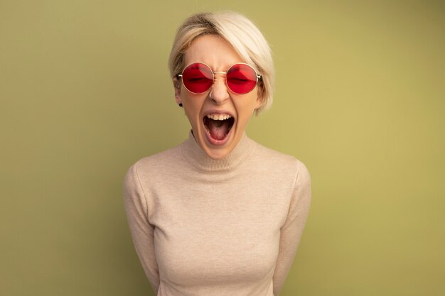 Furiosa joven rubia con gafas de sol manteniendo las manos detrás de la espalda mirando gritando aislado en la pared verde oliva con espacio de copia