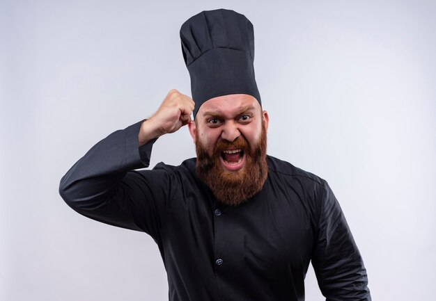 Un furioso chef barbudo con uniforme negro levantando el puño cerrado mientras mira en una pared blanca
