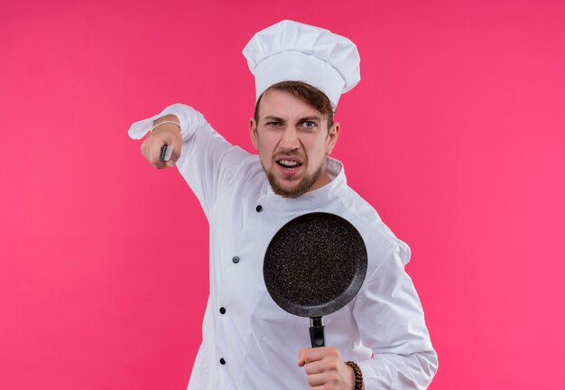 Un furioso joven chef barbudo con uniforme blanco con gorro de cocinero sosteniendo una sartén con un cuchillo mientras mira con expresión negativa en una pared rosa