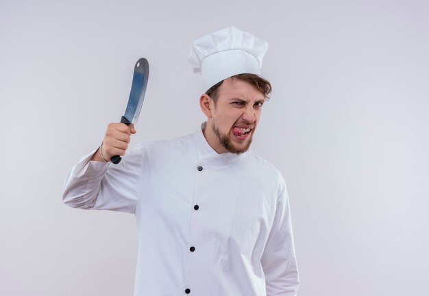 Un furioso joven chef barbudo con uniforme blanco sosteniendo un cuchillo de carnicero mientras mira en una pared blanca