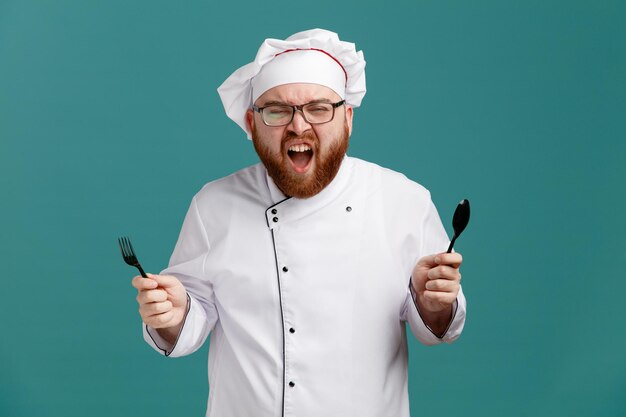 Furioso joven chef masculino con uniforme de anteojos y gorra sosteniendo cuchara y tenedor mirando a la cámara gritando aislado en el fondo azul