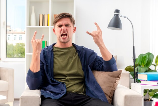 Furioso joven guapo rubio se sienta en un sillón gesticulando cuernos signo de mano con las dos manos